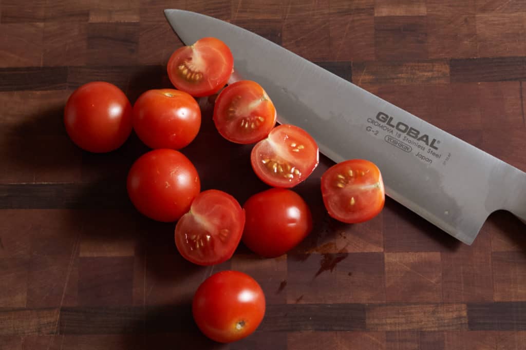 A chef's knife on a cutting board with sliced cherry tomatoes.