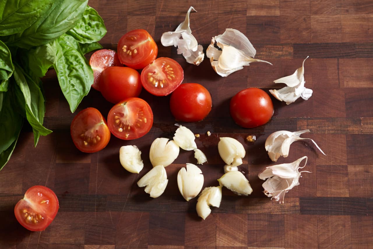 Fresh basil, sliced cherry tomatoes, and crushed garlic cloves on a cutting board.