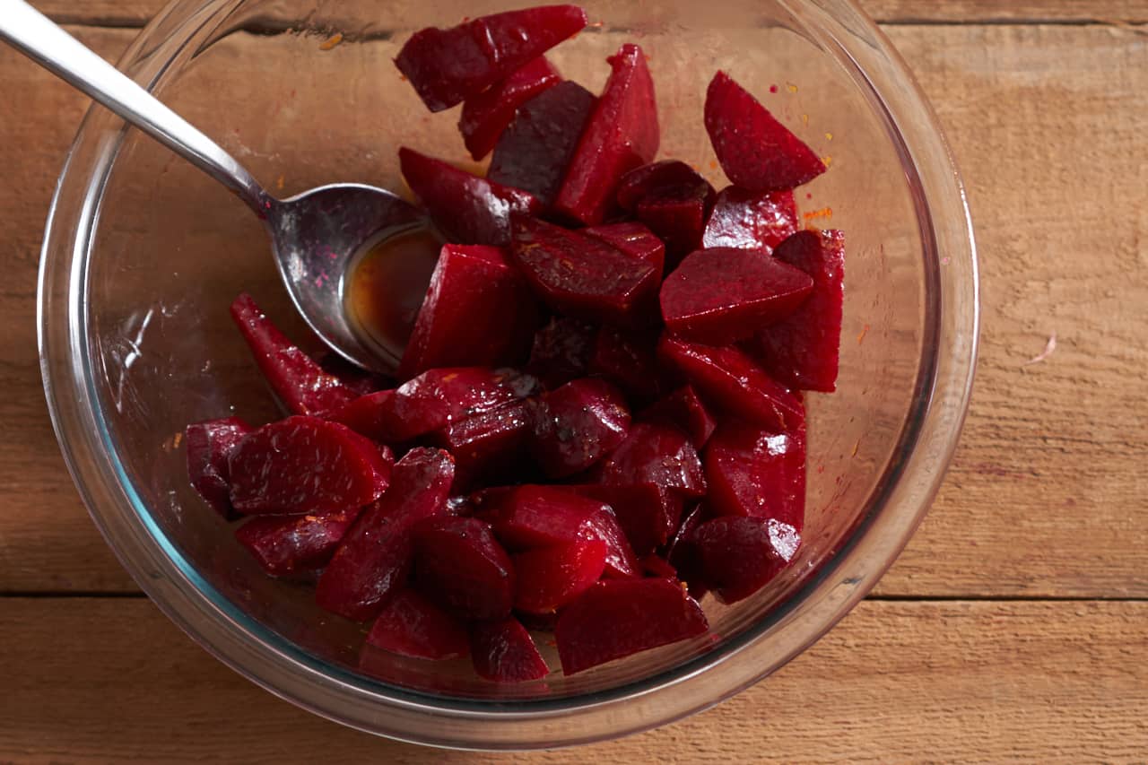 A spoon rests in a glass bowl filled with roasted beets sliced into wedges tossed in orange vinaigrette.