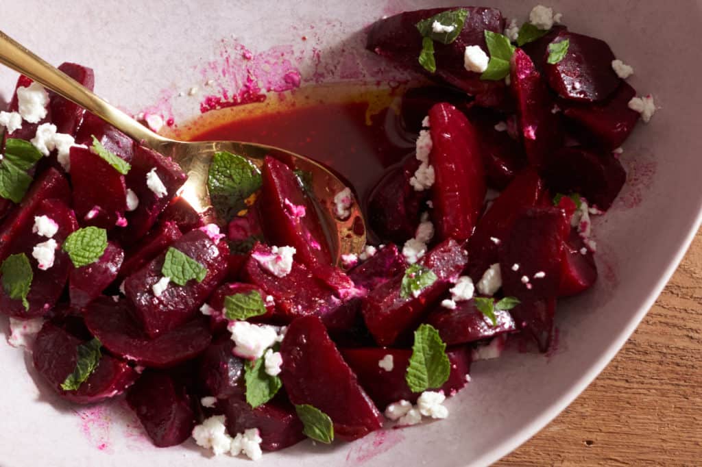 A gold spoon sits in a pink bowl full of roasted beet salad with goat cheese, orange vinaigrette, and mint.