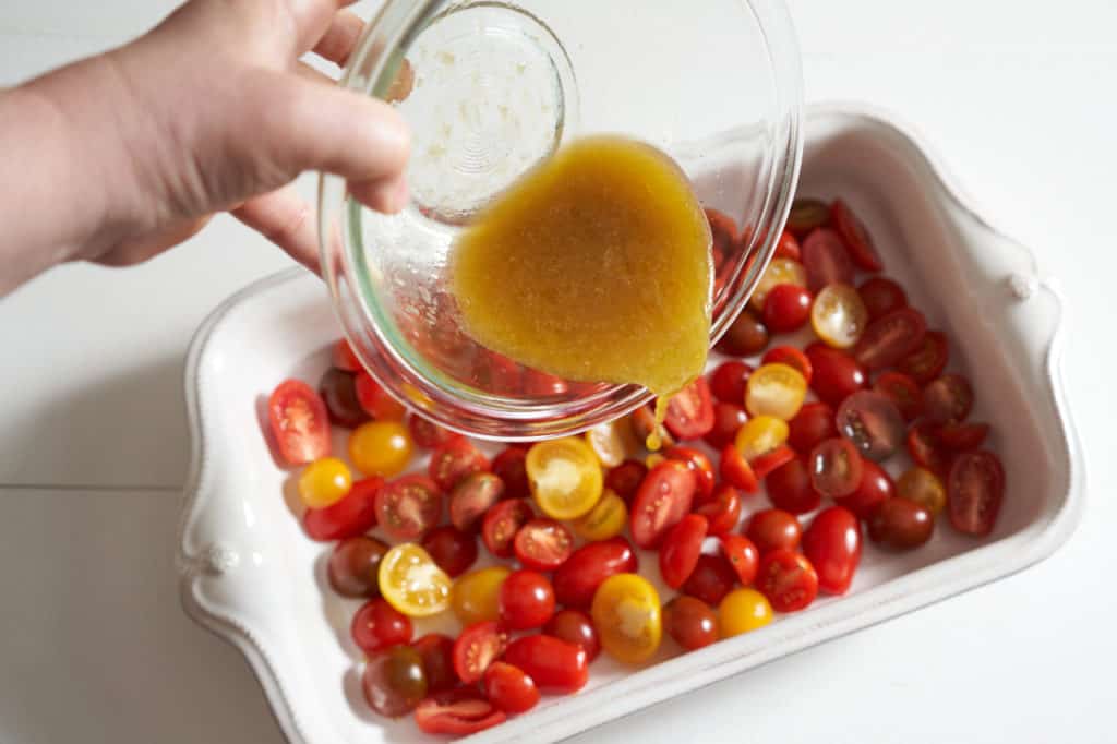A woman's hand is shown pouring a glass bowl of sauce over sliced cherry tomatoes.