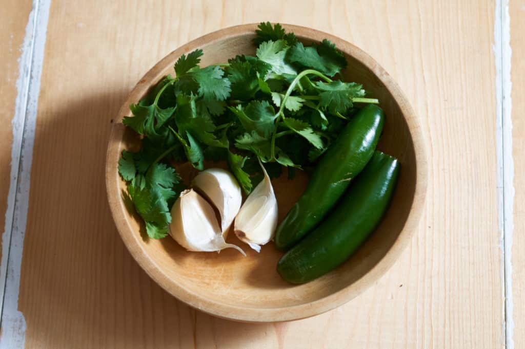 Two serrano peppers, fresh cilantro and three garlic cloves in a wooden bowl.