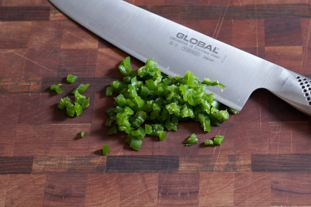 A chef's knife on a cutting board with diced serrano chile peppers.