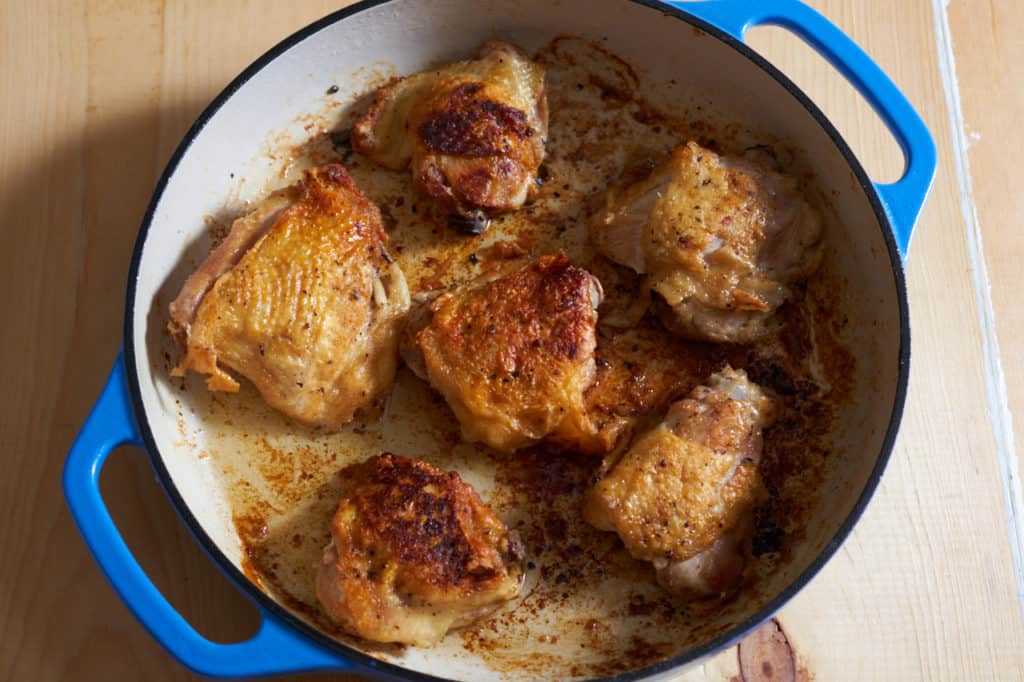 Six browned chicken thighs in a blue casserole pan.