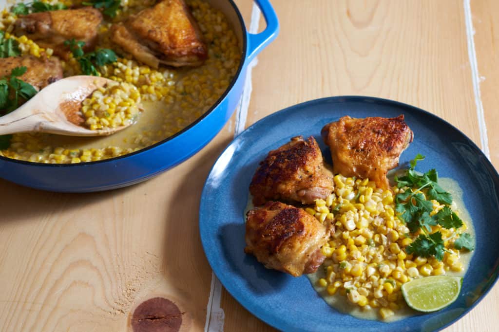 Chicken thighs with coconut milk creamed corn on a blue plate garnished with lime and cilantro, next to a blue casserole pan with more chicken and corn.