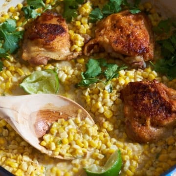 A wooden spoon in a pan of chicken thighs with coconut milk creamed corn, garnished with cilantro and lime wedges.