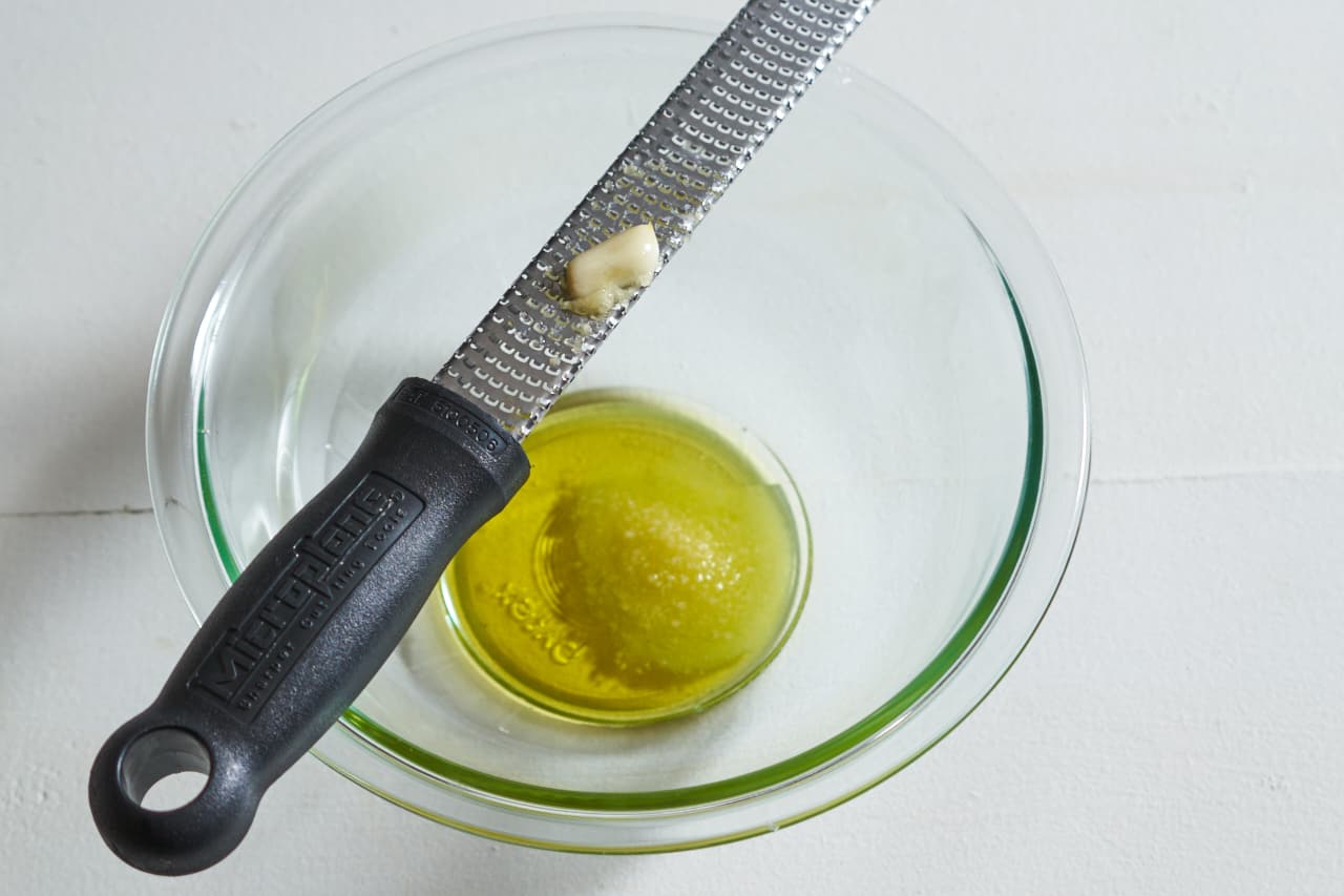 A microplane grater with garlic sits atop a glass bowl filled with olive oil and salt.