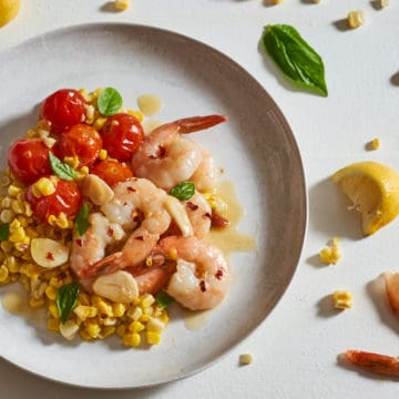 A plate of shrimp scampi with tomatoes and corn surrounded by corn kernels, shrimp tails and basil leaves.