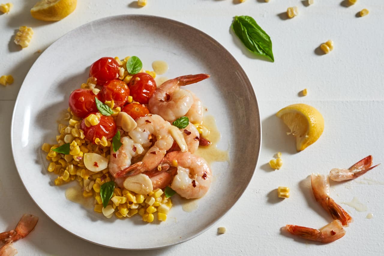 A plate of shrimp scampi with tomatoes and corn surrounded by corn kernels, shrimp tails and basil leaves.