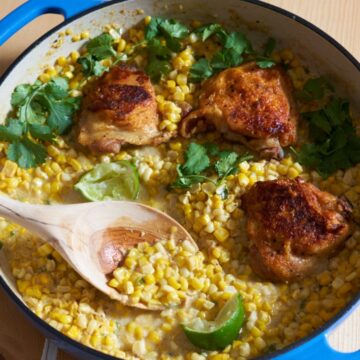 Chicken thighs with coconut milk and corn topped with fresh cilantro and lime wedges in a blue dish with a wooden spoon in it.