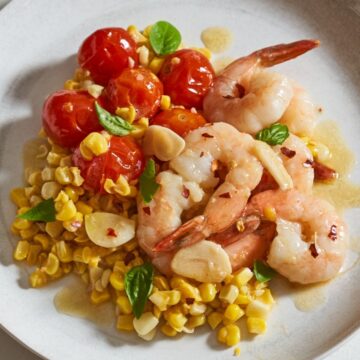 Shrimp scampi served over tomatoes and corn, topped with fresh basil on a white plate.