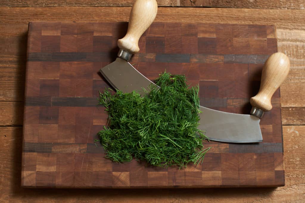 A mezzaluna with chopped dill on a cutting board.