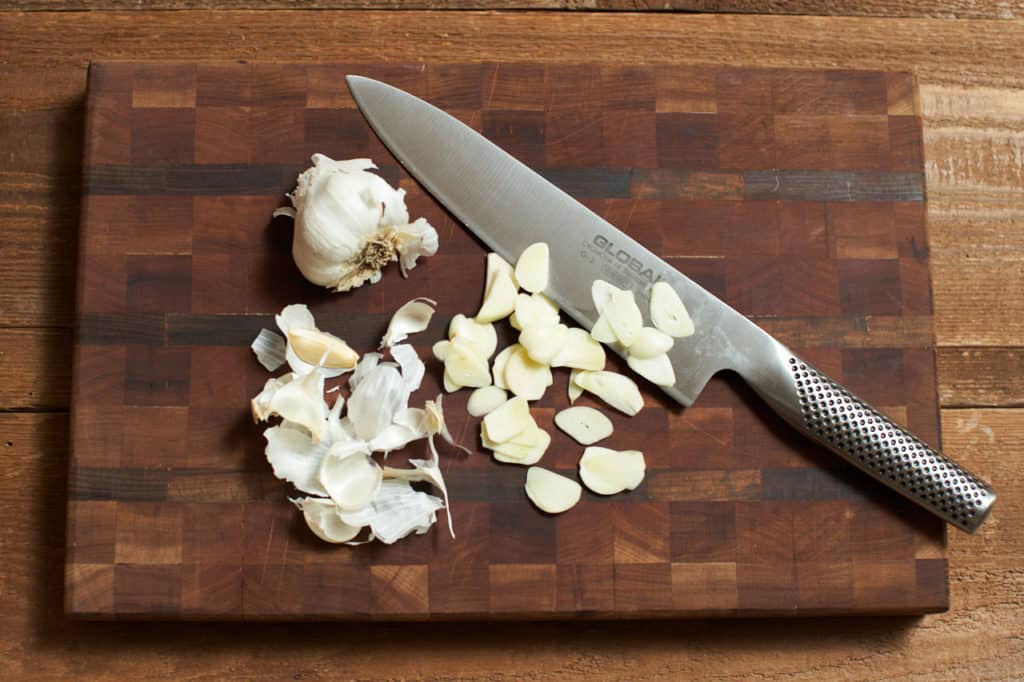 A chef's knife with sliced garlic on a wooden cutting board.