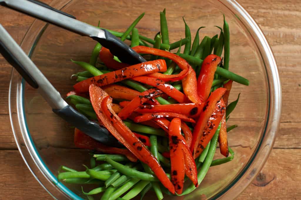 Kitchen tongs sit in a glass bowl with green beans (haricots verts) and charred red peppers.