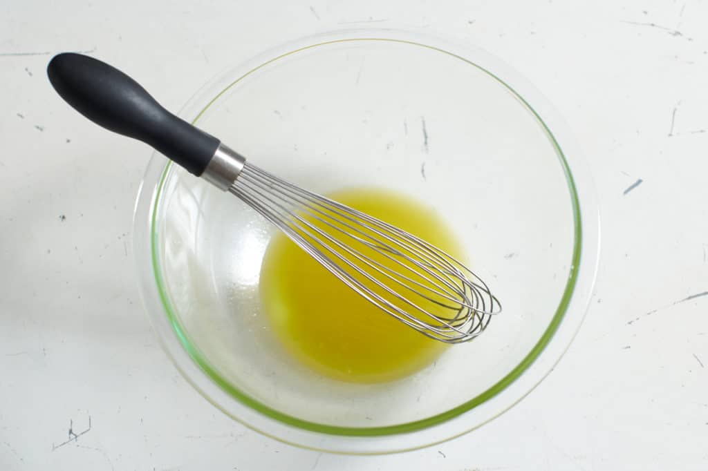 A whisk sits in a glass bowl with olive oil.