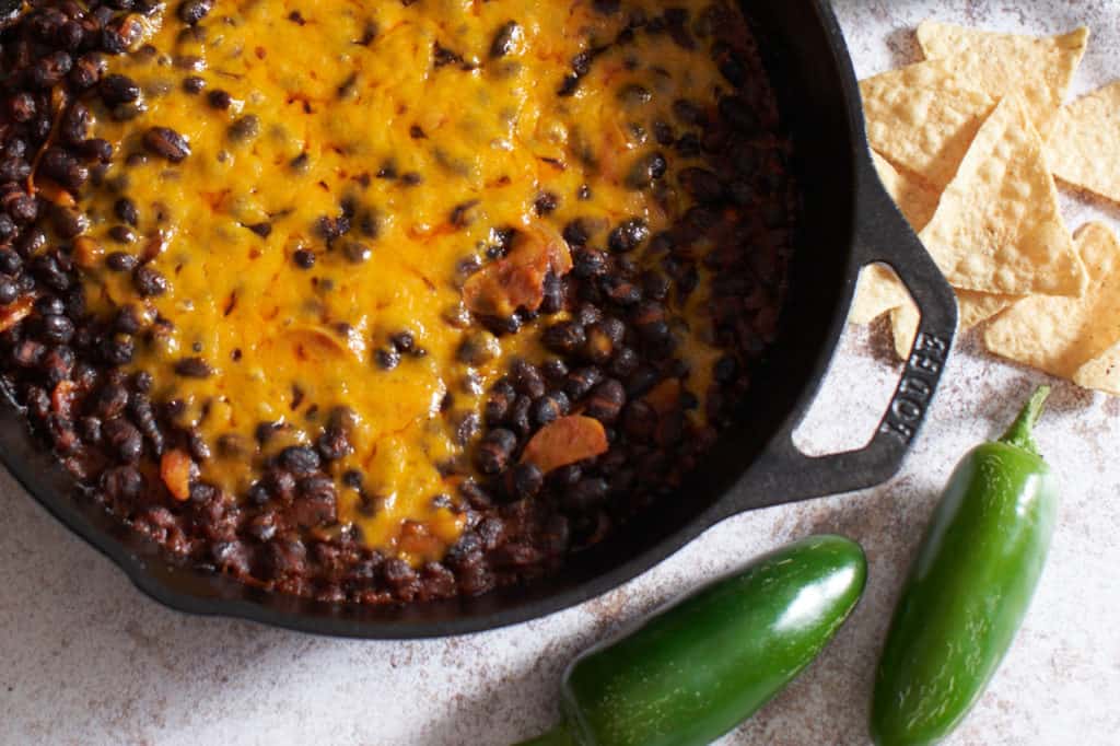 Spicy black beans with melted cheddar cheese in a cast iron skillet, with tortilla chips and jalapeños.