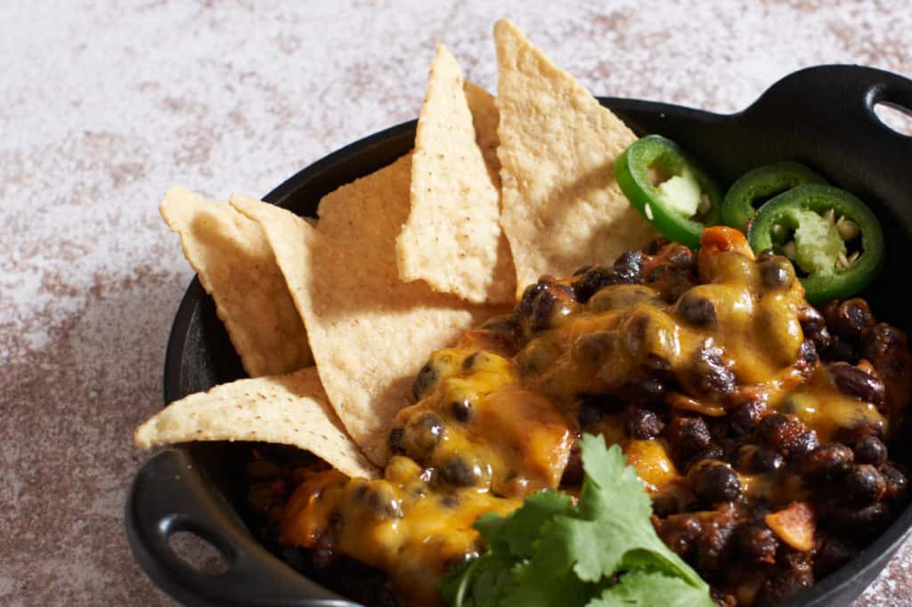 Spicy black beans with melted cheddar cheese, tortilla chips, jalapeños and cilantro in a small cast-iron dish.
