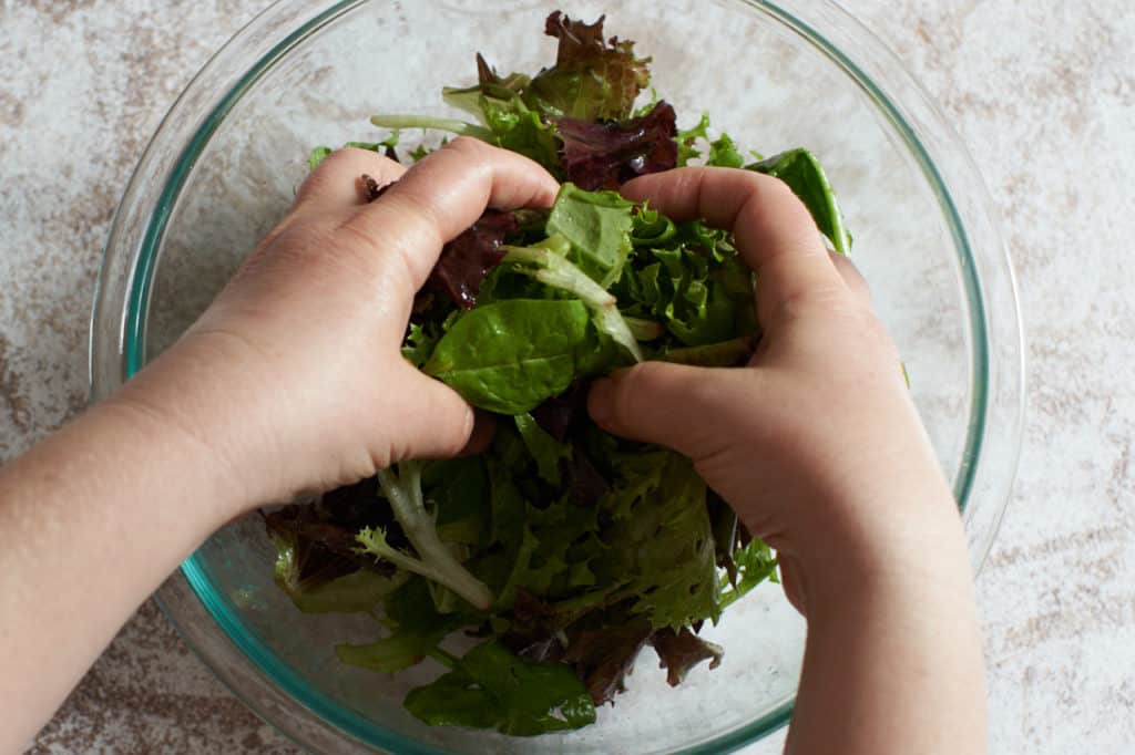 A woman's hands are shown mixing salad greens in a glass bowl.