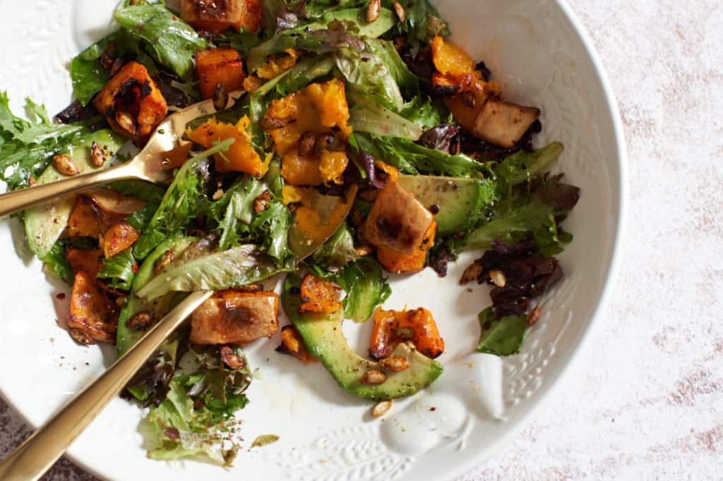 A bowl of harissa roasted butternut squash salad with avocado with gold utensils in it.