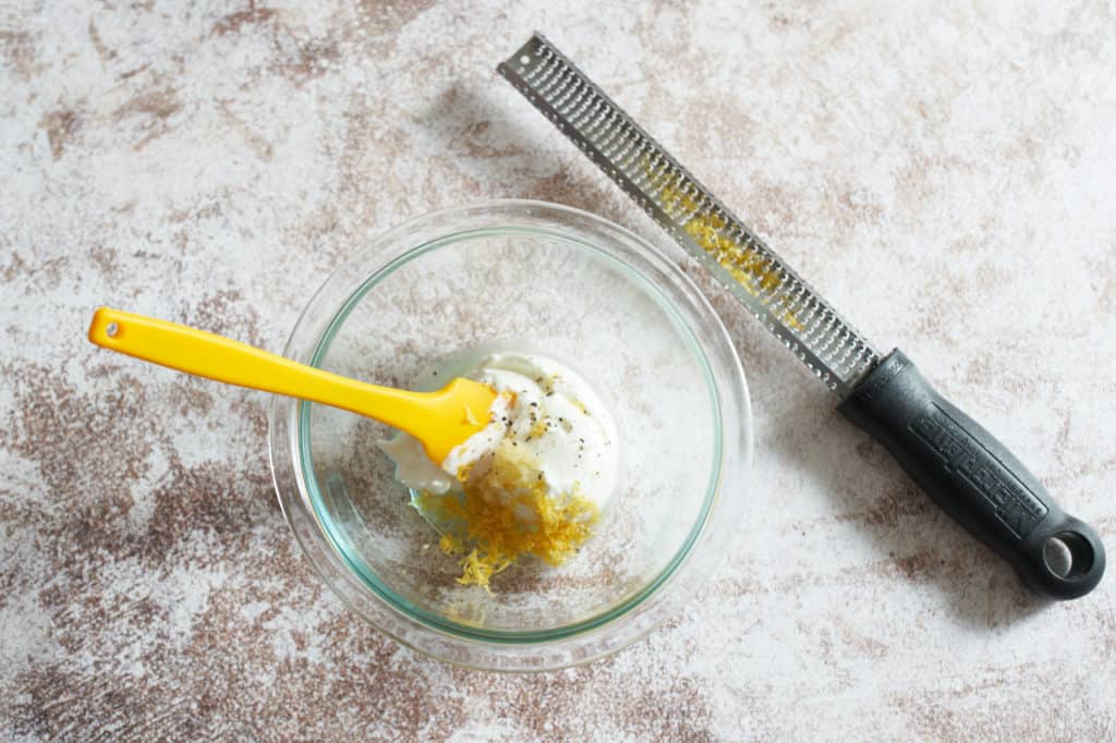 A bowl of yogurt and lemon zest with a yellow spatula in it, and a microplane grater beside it.