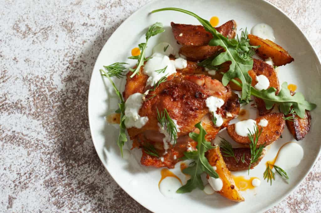 Harissa chicken thighs and potatoes on a white plate topped with herbs and yogurt sauce.