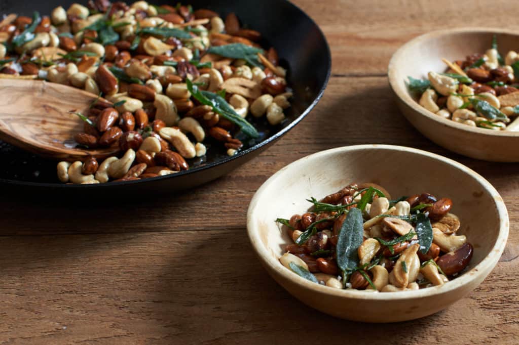 oven roasted nuts with herbs and garlic in a skillet next to two small wooden bowls filled with nuts