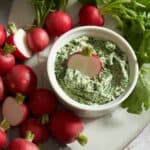 Fresh radishes on a platter surrounding a crock of radish leaf butter with half a radish dipped into it.