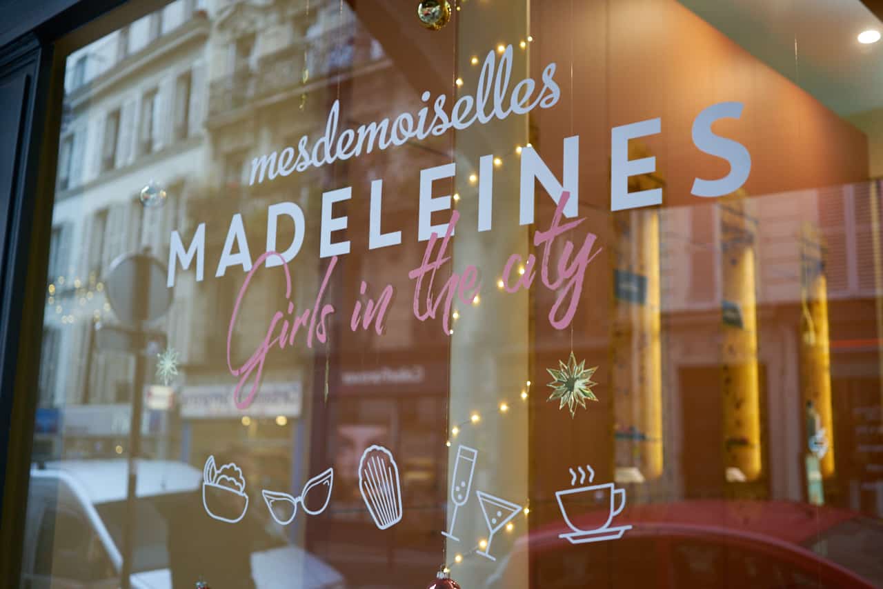 The window of a madeleine bakery in Paris, France. White and pink writing says, "Mesdemoiselles Madeleines, Girls in the City" with drawings of beverages, sunglasses and madeleines underneath.