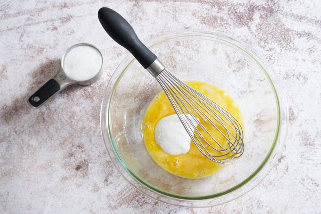 A whisk in a glass bowl with beaten eggs and sugar.