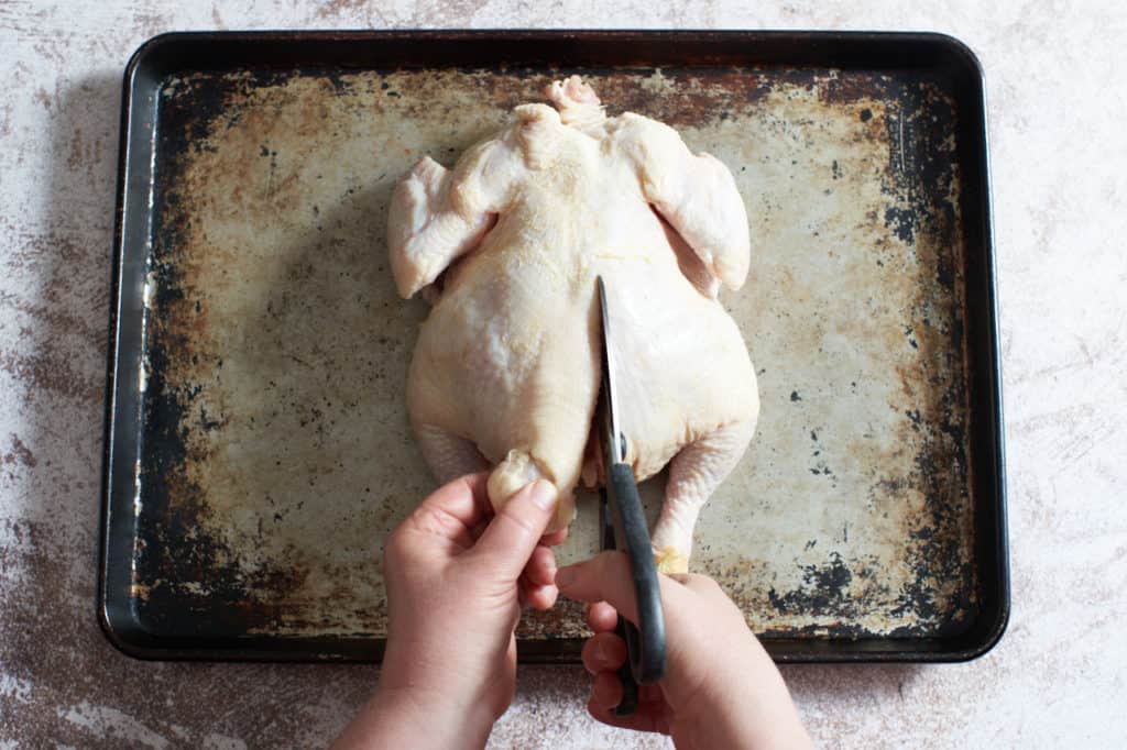 A woman's hands cutting the backbone out of a chicken with kitchen shears.
