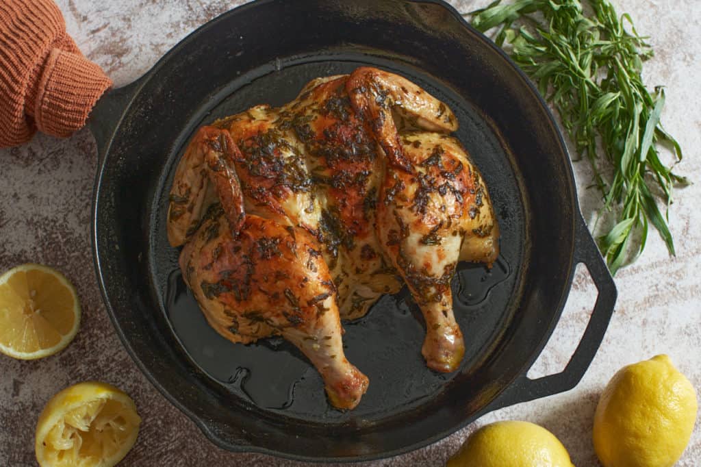 A roast chicken with tarragon in a cast iron skillet surrounded by fresh lemons and fresh tarragon. 