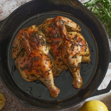 A roast chicken with tarragon in a cast iron skillet, surrounded by lemons and fresh tarragon.
