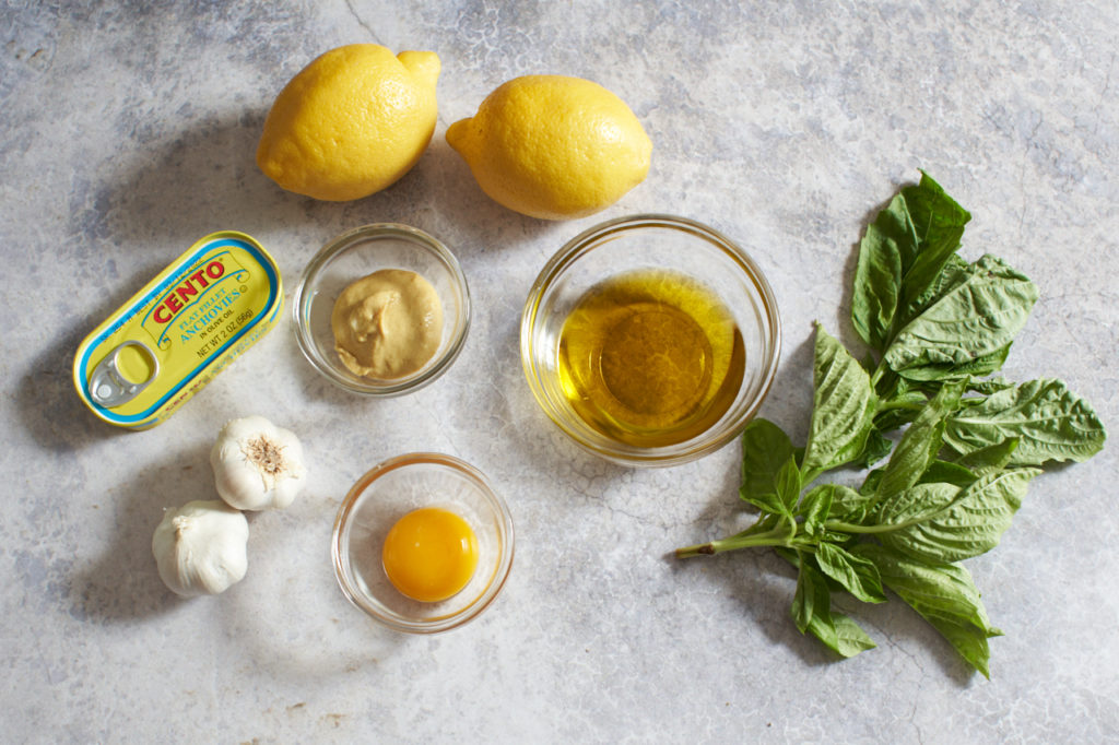A can of anchovies, lemons, garlic, fresh basil, and three small bowls filled with olive oil, mustard, and an egg yolk.