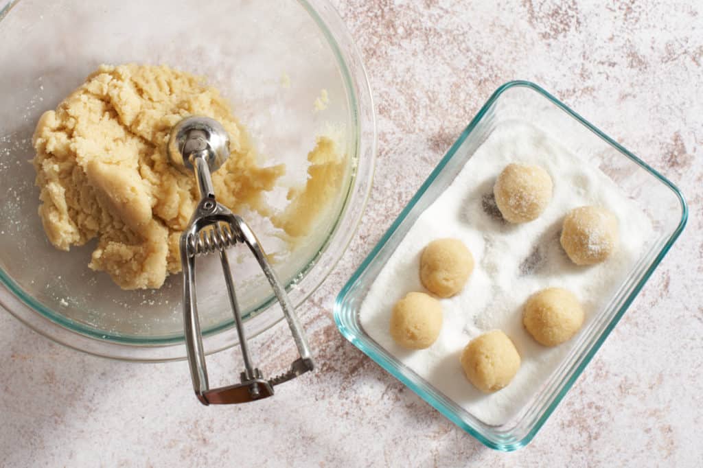 A bowl of cookie dough with a scoop and a glass tray of cookies rolled in sugar.