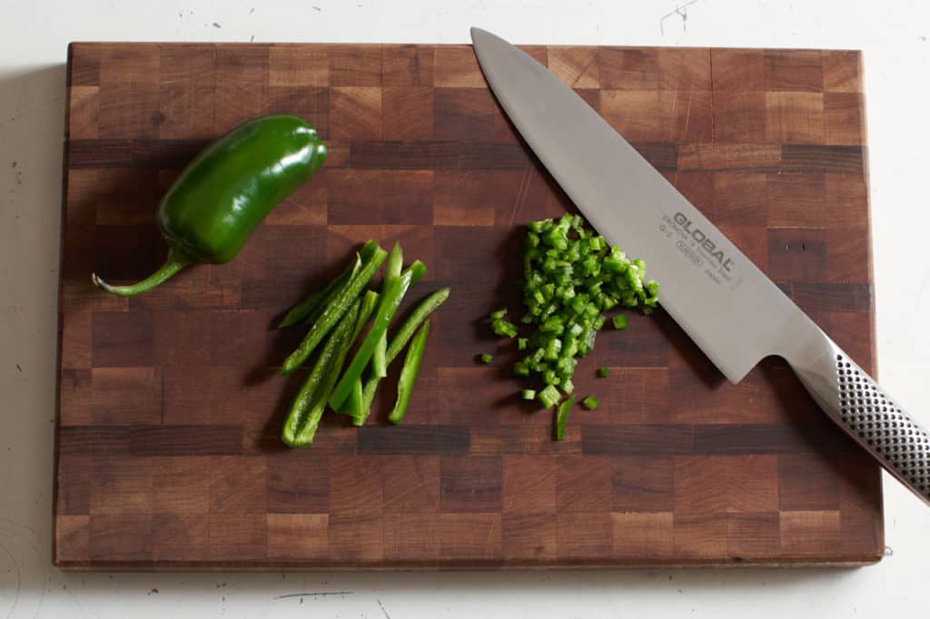 A knife rests on a cutting board with chopped jalapeños.Some are sliced into strips and some are diced.