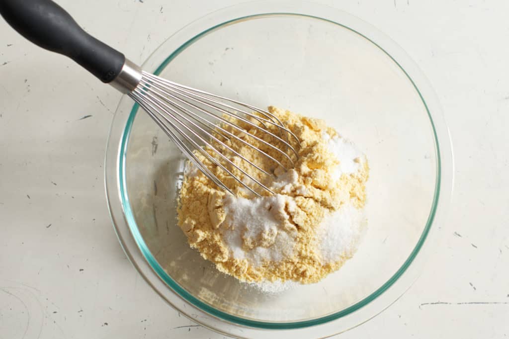 A whisk in a glass bowl of cornmeal with sugar and baking powder.