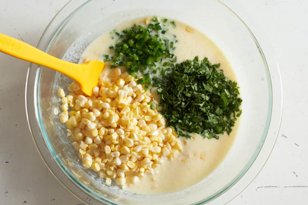 A yellow spatula in a glass bowl with batter and fresh corn, jalapeños and cilantro.