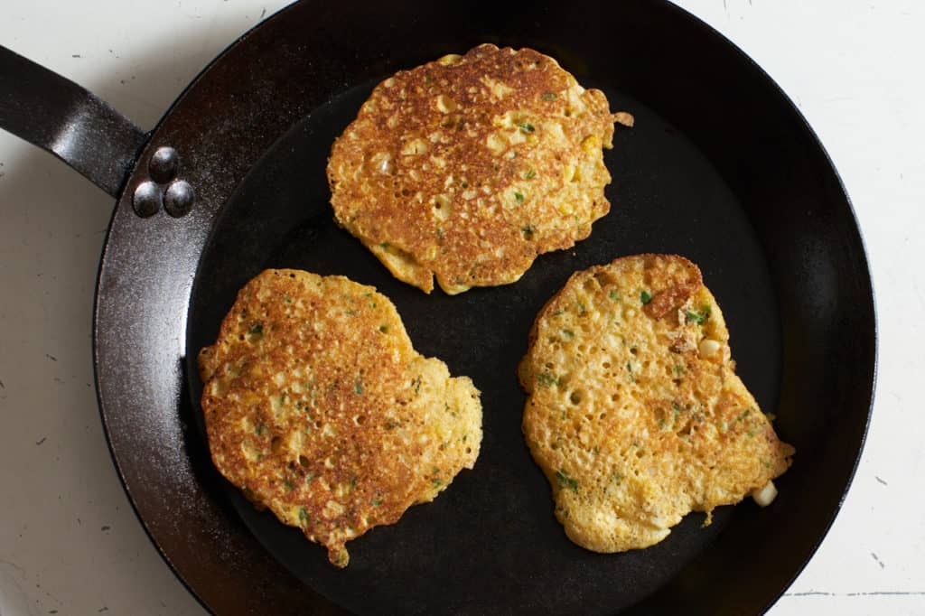 Three golden brown corn cakes in a carbon steel skillet.