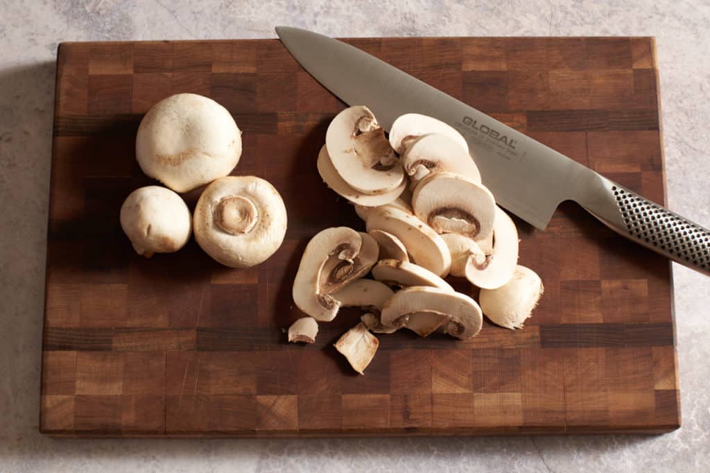 A knife on a cutting board with sliced and whole mushrooms.
