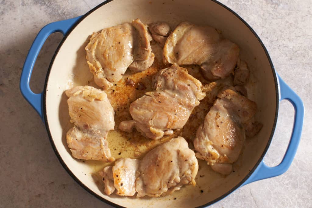 Browned chicken thighs in a blue casserole pan.