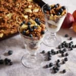Two yogurt parfaits on a marble surface with blueberries and peaches and granola in the background..