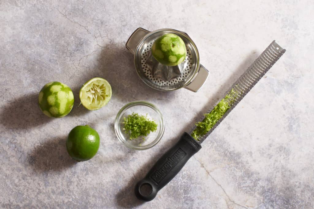 A grater with lime zest, a small manual juicer with half a lime, and fresh limes.