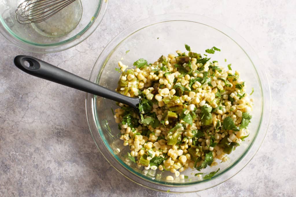 A black spatula in a glass bowl of corn salad tossed with lime dressing.