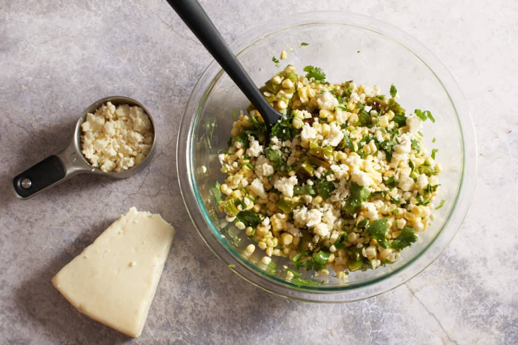 A black spatula in a bowl full of corn salad next to a wedge of cotija cheese and a measuring cup full of cheese.