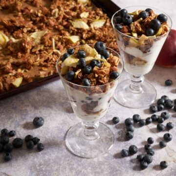 Peach blueberry yogurt parfaits in tall glasses in front of of a sheet tray of granola, surrounded by fresh blueberries.