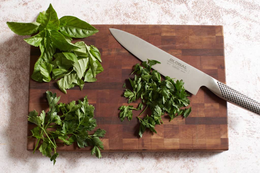 A knife on a cutting board with chopped herbs.