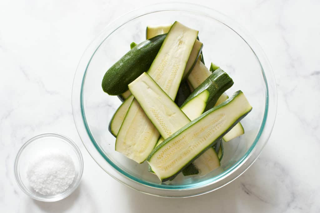 Sliced zucchini in a glass bowl with a small bowl of salt next to it.