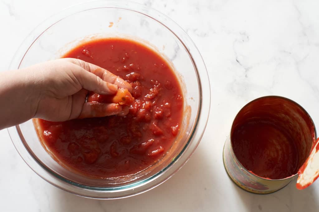 A woman's hand in a bowl of crushed tomatoes with an empty can to the right.