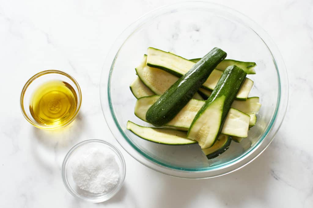 A bowl of sliced zucchini with two small bowls with olive oil and salt to the left.