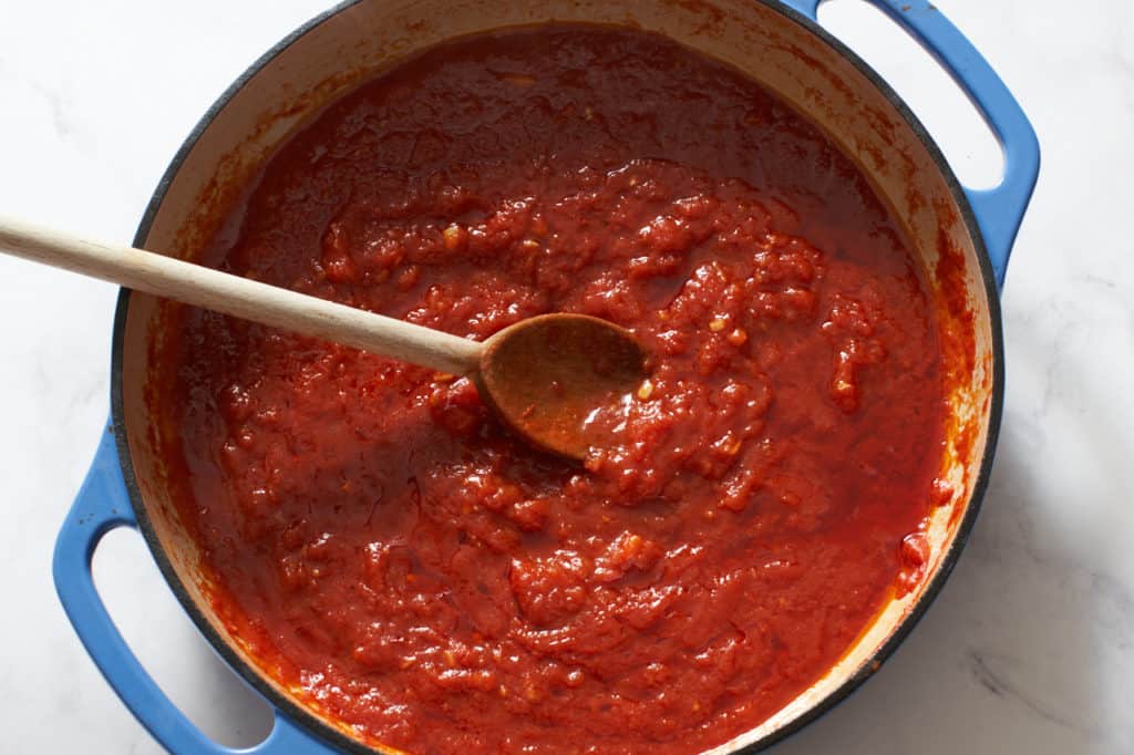 A wooden spoon in a blue casserole pan with tomato sauce.
